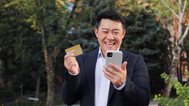 現代のスマートフォンやスポーツベットゲームを探してクレジットカードを保持している男性の手のクローズアップ 中国の幸せなビジネスマンは街の通りに立って オンラインショッピングを楽しんで 満足した買い手笑顔 — ストック動画