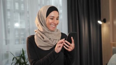 Güzel, gülümseyen, başörtüsü takmış Müslüman bir kadın evde akıllı telefon kullanıyor. Modern dairesinde elinde cep telefonu olan orta doğulu bir kızın portresini yakından çekiyor.
