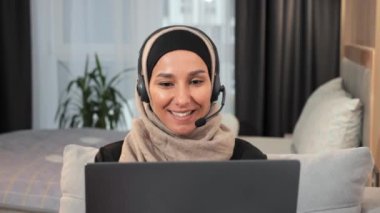 Güzel, genç, Arap Müslüman kadın blogcu, kulaklık takmış evde oturmuş bilgisayarla internetten video görüşmesi yapıyor. Türbanlı kadın vlogger çalışıyor, uzak kurslar. Video blogu. Müslümanlar. Eğitim