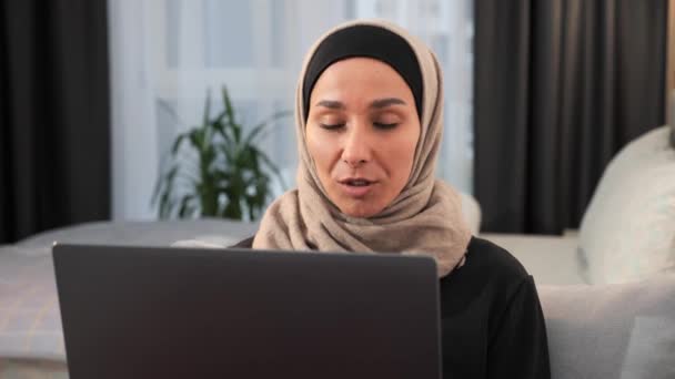 自信を持ってアジアのイスラム教徒の女性のヒジャーブでは ラップトップのウェブカメラに話してビデオ通話を行います イスラムビジネスの女性は 自宅のオフィスでチームとオンライン会議を作業し ビデオ通話を議論している — ストック動画