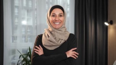 Ev ofisinin arka planında geleneksel başörtüsü takarak kameraya gülümseyen profesyonel müslüman iş kadınının yakın portresi. Güzel bir kadın İslami yüz, kapalı mekanda eller.