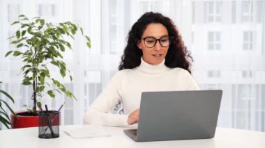 Gözlüklü Latin kökenli genç bir iş kadını ev telefonuyla iletişim kuruyor. Etnik iş kadını dizüstü bilgisayara bakıyor, çevrimiçi öğrenim konferansı, sanal eğitim konsepti