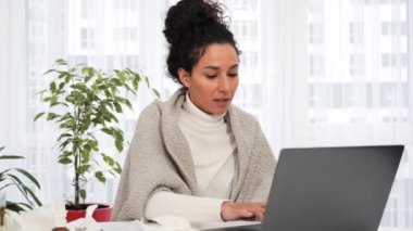 Laptop bilgisayarında çalışan, öksüren ve hapşıran sağlıksız, hasta, genç bir Latin kadının yüksek ateşi, soğuk algınlığı ve virüs enfeksiyonu var. Hasta kadın grip olmuş, battaniyeye sarılmış, yakalanmış.