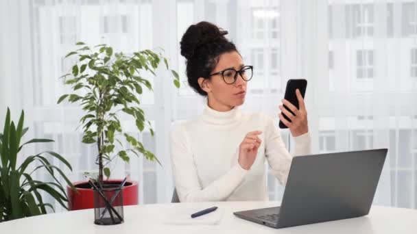 自宅でテーブルの上に座っている白いセーターと眼鏡の遭難若いラテン語の女性は 彼女のスマートフォン上のセルラー接続やインターネットがない理由を理解していません 通信の問題 — ストック動画