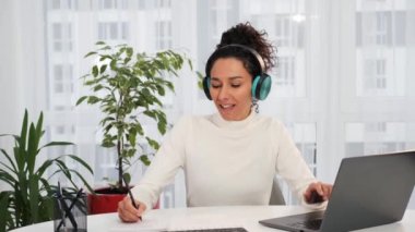 Aşırı mutlu genç Latin iş kadını kablosuz kulaklık takıyor akıllı telefon dansında müzik dinliyor, gülümsüyor, kaliteli bir ses çıkarıyor ev işi masasında el sallayarak sandalyede oturuyor.