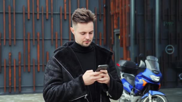 青いオートバイの近くに屋外に立って冬のジャケットを着て成功したビジネスマンのフリーランスの笑顔の肖像画 携帯電話にスマホスクロール入力メッセージを保持している男 ライフスタイルコンセプト — ストック動画