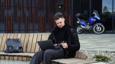 Sırt çantalı genç adam bankta oturuyor, elinde kredi kartı var. Güvenlik kodunu gir, online alışveriş yap, uzaktan çalış, e-ticaret yap.