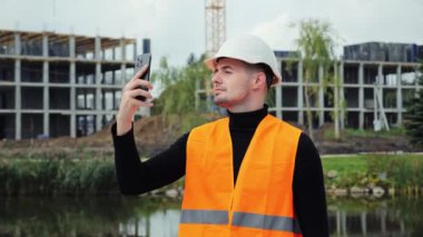 Beyaz kasklı çekici bir mühendis cep telefonuyla inşaat halindeki binaların fotoğraflarını çekiyor. Erkek kişi akıllı telefondan müşteriye mesaj gönderir. Mühendislik projesi