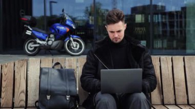 Sıcak moda kıyafetleri giyen genç adam dizüstü bilgisayarda çalışıyor sırt çantasıyla bankta oturuyor mavi motosiklet arka planında dinleniyor mobil ofis serbest iş hayatı konsepti