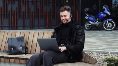 Yakışıklı bir iş adamı. Mavi bir motosikletin arka planında kulaklık ve dizüstü bilgisayarla uzaktan yakından çalışıyor. Video konferans mesafesi toplantısı. İş adamı internette sohbet ediyor.