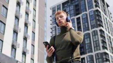 Kulaklık takan neşeli moda adamı yaz günü modern şehirde akıllı telefon müzik uygulaması kullanarak dışarıda dans ediyor. Yakışıklı adam eğleniyor, şehir sokağında dinleniyor. İnsanlar ve müzik konsepti