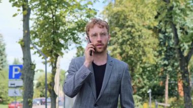 Güzel taranmış saç stili ve akıllı telefondan konuşan sakallı çekici bir adam. Hoş ve ilginç bir görünüş. Yakışıklı adam açık havada yürürken aktif olarak cep telefonuyla konuşuyor.