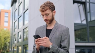 Sosyal medyada Genç Mutlu İş Adamı Mesajlaşması 'nın kapanışı. Akıllı Telefon Ekranı 'na bakıyorum. Klasik giyiniklik. Arka plandaki Büyük Cam Bina. Başarılı ve kendinden emin görünüyor.