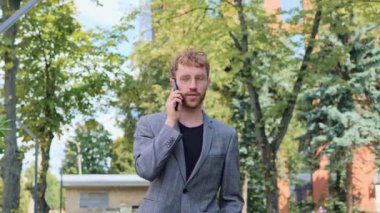 Havalı stil sahibi iş adamı dışarıda cep telefonuyla konuşuyor. Gözlüklü neşeli yakışıklı adam iş yerinde ağır çekimde telefonla konuşuyor. Kendine güvenen iş adamının portresi dışarıdaki telefonu arıyor.