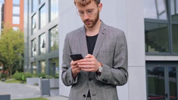 在社交媒体上 穿着时髦紧身衣 戴着眼镜的时尚小伙通过手机应用发邮件给年轻快乐商人发短信 看他的智能手机屏幕 — 图库视频影像