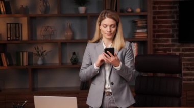 Resmi takım elbise giyen zarif mutlu iş kadını mobil zaferi kutluyor, şanslı yönetici, akıllı telefondan iyi haberler alıyor, dahili ofiste mali başarı iddiasına seviniyor.