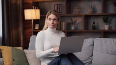 Mutlu genç bir kadın elinde dizüstü bilgisayarla kanepede oturup internette sörf yapıyor sosyal medya uygulamaları ya da uzak mesafe işleri kullanarak internetten alışveriş yapıyor ve evdeki modern teknoloji aletleri üzerine eğitim görüyor.