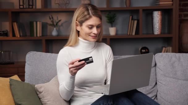 かなり幸せな女の子は自宅でソファに座って笑顔で現代のラップトップを使用してクレジットカードを保持するオンライン決済を行っています インターネットバンキングのショッピング 電子商取引の概念 — ストック動画