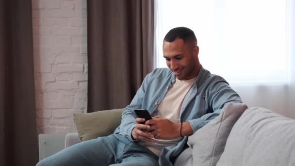ソファに座っている間にスマートフォンを使っている陽気な若いアフリカ人男性 アフリカ系アメリカ人の笑顔の男自宅で携帯電話のアプリを使用しています ハンサムな男性リラックスチャット上の携帯電話スタンド屋内 — ストック動画