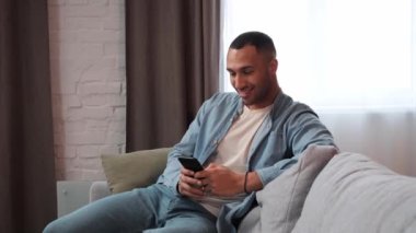 Evdeki rahat oturma odasında akıllı telefon kullanan yakışıklı Afrikalı Amerikalı adam. Adam rahat koltukta dinleniyor. İnterneti tarıyor ve sosyal ağlarda videoları kontrol ediyor ve eğleniyor.