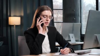 Ofis içinde dizüstü bilgisayarla çalışan odaklanmış bir iş kadını. Kadın işçi telefon görüşmesi yapıyor. Uzaktan kumandalı iş yerinde akıllı telefondan konuşan bir kadın. İş kadını modern ofisten telefon ediyor.
