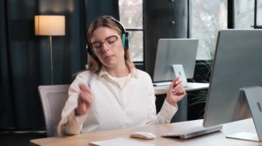Amusing business woman manager worker sits at table in headphones, listens music, makes wave with hands, dances in corporate office drinks coffee. Comic young cheerful girl relaxes to favourite song