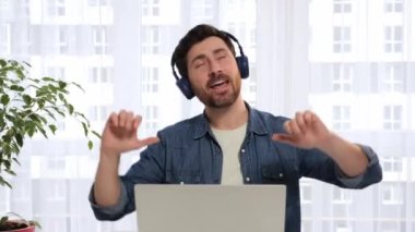 Funny business man manager worker freelancer sits at table in headphones, listens music, makes wave with hands, dances having fun, work from home. Comic young cheerful man relaxes to favourite song