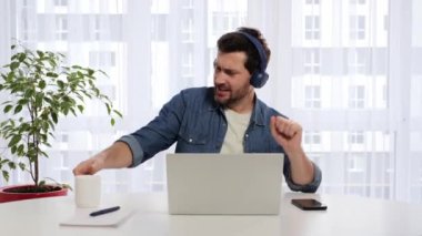 Active young man freelancer manager sits at desk in front of laptop wearing headphones, dances, moves waves arms, listens to music, feels joy enjoyment of freedom, relaxes on break, funny movements