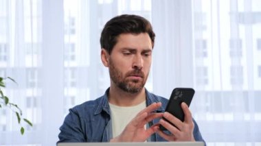 Shocked man sitting at home kitchen reading terrible news on smartphone. Upset business man looking with horror at mobile phone and touching to head with hand. Unpleasantly surprised reacting message