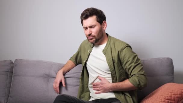 胃の中にひどい痛み 腹部の痛みに苦しんでいるカジュアルな服を着た若い男が 自宅のソファの上に座っている 男は突然強い胃の痛み 胃炎の問題を感じる — ストック動画