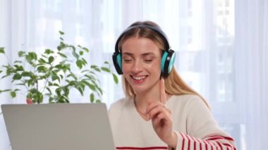 Happy businesswoman manager worker freelancer sits at table in headphones, listens music, makes wave with hands, dances having fun, work from home. Comic young cheerful girl relaxes to favourite song