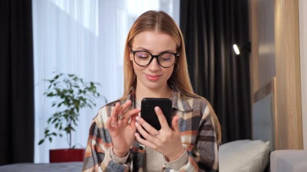 自宅でスマートフォンの食事を使用して スマートカジュアル服や眼鏡を身に着けている愛らしい美しいブロンドの女性のフリーランサー マネージャーオンライン人々との接続 ブラウジングインターネット ソーシャルメディア — ストック動画