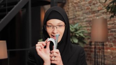 Rich Muslim woman in hijab and glasses smiling with cash make money dollar waving fan posing at modern office. Portrait happy successful business freelancer female with currency stack salary income