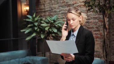 Professional female businesswoman worker having mobile phone call with the customer doing paperwork standing in office. Young leader woman holding document working on project making business call
