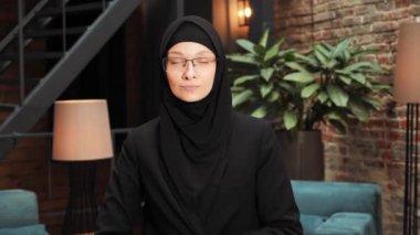 Closeup focused arabian businesswoman in eyeglasses raising head smiling to camera. Muslim female face portrait islam arab girl happy pretty islamic woman wears black hijab standing in meeting room