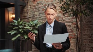Confident professional businesswoman worker looking at chart while writing typing message or note on mobile phone standing in office. Young leader Woman holding a document working on a project