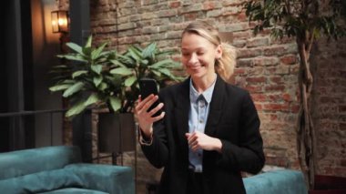 Smiling business woman having video call on smartphone in a modern office room or conference hall. Positive female manager making video conference chat online on mobile phone. Remote work