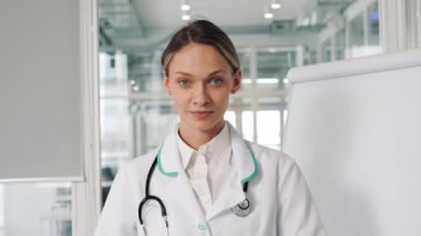 Confident young european woman doctor wearing white medical coat and stethoscope looking at camera in clinic. Smiling female physician posing in hospital office. Positive practitioner face portrait