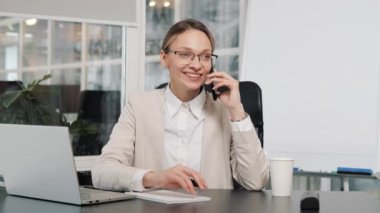 Smiling confident businesswoman in glasses talking calling on mobile phone in office having friedly conversation Cheerful happy manager female CEO woman using smartphone writing notes gesturing hands