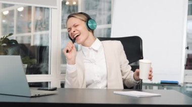 Cheerful happy positive adult woman dancing listening music in headphones at workplace sing song to pen like microphone. Business lady businesswoman entrepreneur worker in office funny dance singing