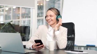 Funny happy young woman in headphones singing holding mobile phone while listening to loud music sitting at office desk with laptop, lazy worker wasting time at workplace in coworking, free rest time