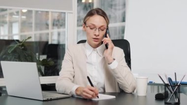 Busy confident businesswoman in glasses talking calling on mobile phone in office having friedly conversation Cheerful happy manager female CEO woman using smartphone writing notes gesturing hands