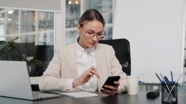 Young busy business woman office worker in eyeglasses working on project paperwork using mobile phone. Smart female manager making budget writing tasks from cellphone working alone in office