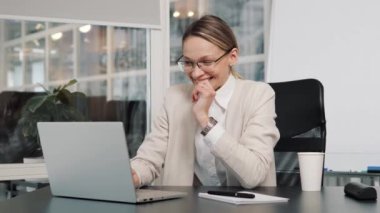 Young business woman manager in glasses and light formal suit sits at table with laptop working from office. Female CEO gets great online news puts hand on chin, satisfied successful entrepreneur