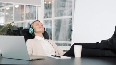 Relaxed happy young woman in headphones singing holding mobile phone while listening music sitting at office desk with laptop, lazy worker wasting time at workplace in coworking, free rest time