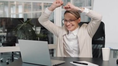 Smiling overjoyed beautiful blonde young woman in light jacket posing doing winner gesture, say Yes i win in office background. Great news celebrating moment of victory, personal achievement