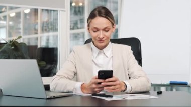 Closeup happy businesswoman using mobile phone tapping message at workplace. Smiling woman browsing internet on smartphone in office. Portrait of business woman reading messages on cellphone