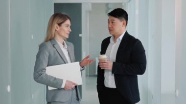 Business woman with laptop in hands and Asian businessman talking in hallway office, talking about work, break diverse colleagues discussing news project in coworking, good relations at workplace