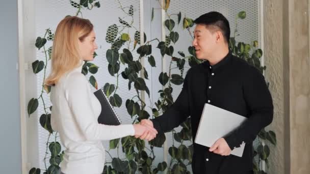 アジアの投資家とヨーロッパのビジネスオーナーの2人の多様なビジネスパートナーがエコオフィサーで緑の植物を背景に握手し 成功した契約を締結します — ストック動画