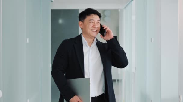 男性ハンサムな韓国のビジネスマンは オフィスホールに立って満面の笑みを浮かべて携帯電話で話しています 優しい会話を持ち ノートパソコンを持ち — ストック動画